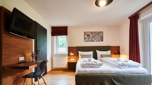 A modern hotel room with a bed, a desk, a chair, a TV, and a window with curtains. - EVI Apartments - Evi (Kaprun)