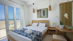 A hotel room with a large bed, a wooden nightstand, a small table with a chair, a wooden wardrobe, and a large window with a view of the beach. - Villa Lavender in Ayia Napa (Sotira)