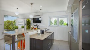 A modern kitchen with a central island, bar stools, a built-in oven, and a large window providing a view of greenery. - Villa Lavender in Ayia Napa (Sotira)