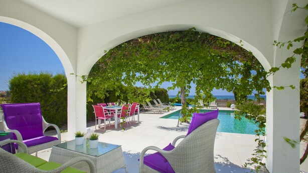 A covered outdoor seating area with white wicker furniture and a glass table, overlooking a pool and the sea. - Villa Lavender in Ayia Napa (Sotira)