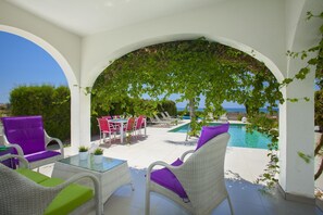 Un área de estar al aire libre cubierta con mobiliario de ratán blanco y una mesa de vidrio, con vistas a una piscina y al mar.