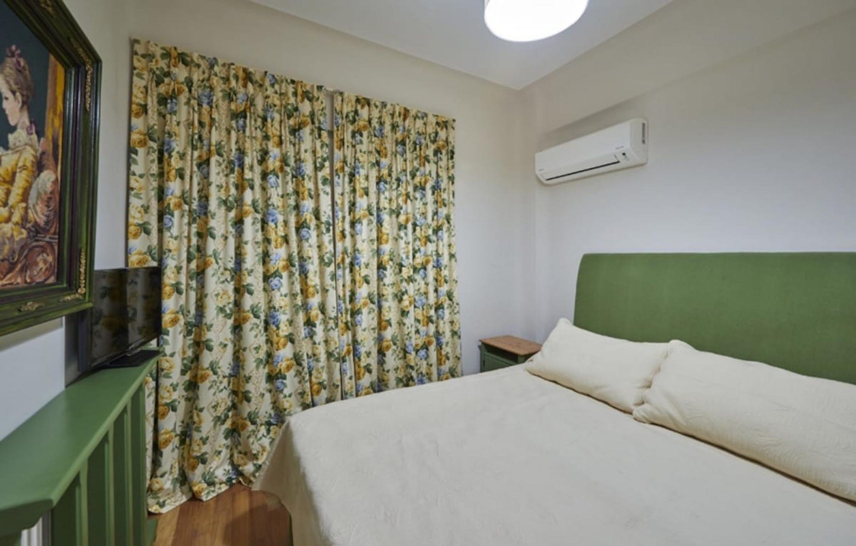 Un dormitorio con cama, cabecera verde, cortina floral, un cuadro en la pared y un equipo de aire acondicionado.