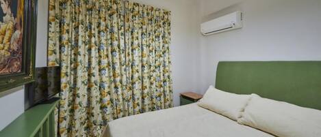 Un dormitorio con cama, cabecera verde, cortina floral, un cuadro en la pared y un equipo de aire acondicionado.