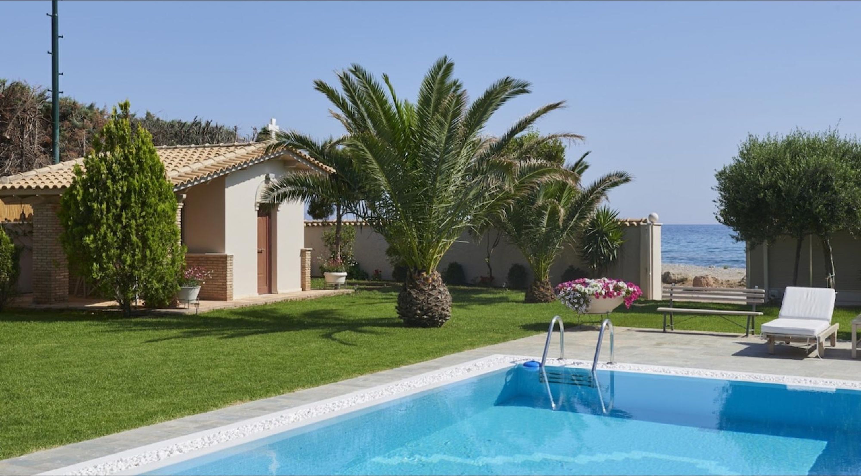 A pool area with a clear view of the sea, a small building, and a palm tree.