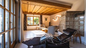 A spacious living room with a leather recliner, a wooden ceiling, a large window with curtains, and a cabinet with glass doors. - Chalet Crown of the Alps (Zell am See)