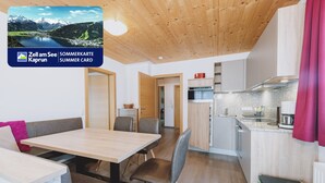 A modern kitchen with wooden cabinets and a countertop, a dining area with a wooden table and chairs, and a view of a mountainous landscape. - Appartements Sulzer - TOP 21 (Zell am See)