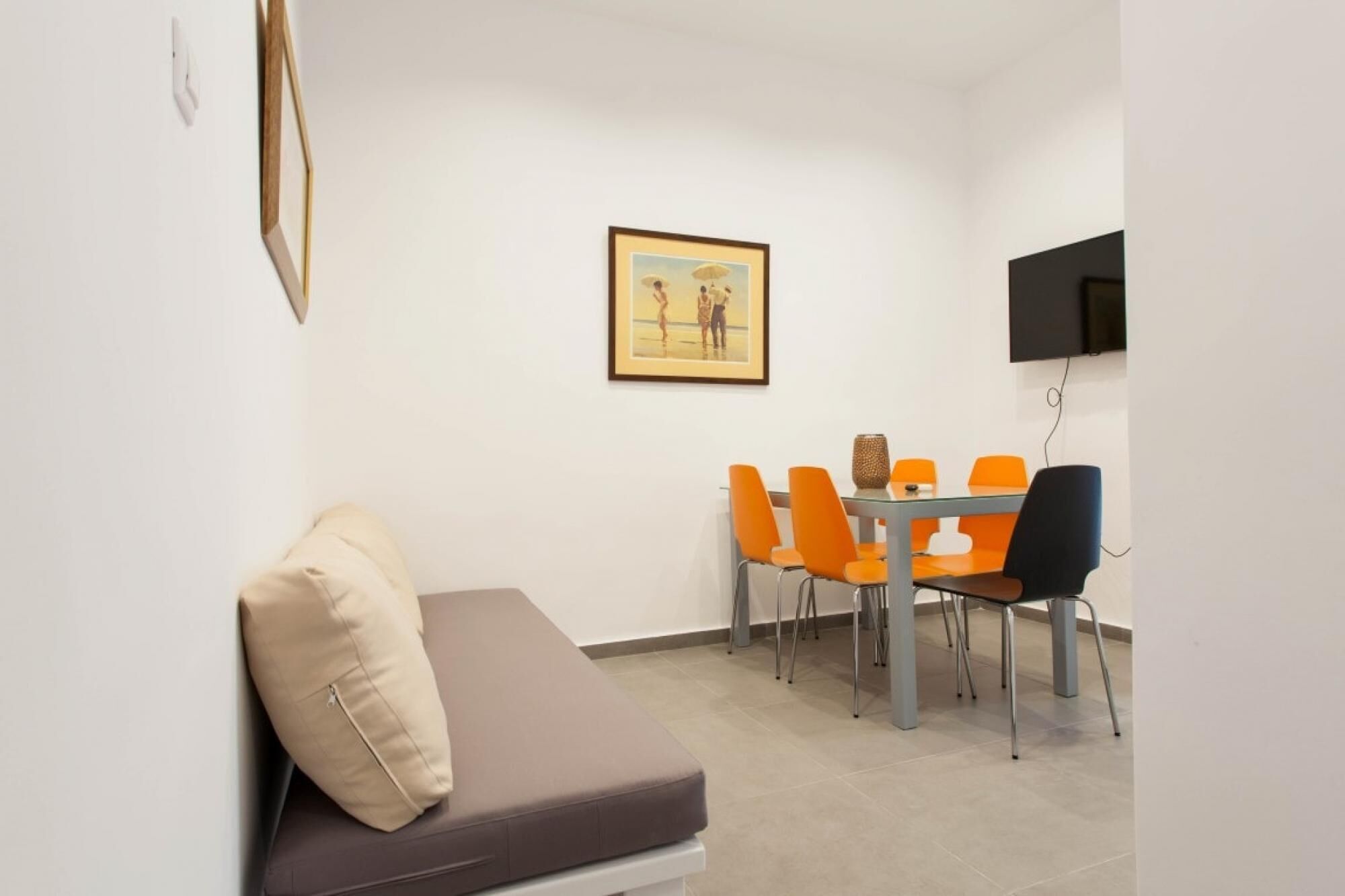 A small, modern room with a sofa, a dining table with orange chairs, a framed picture on the wall, and a flat-screen TV mounted above the table.