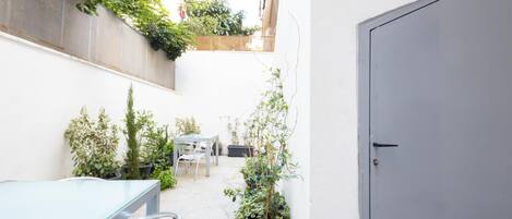 A narrow courtyard with a glass table, potted plants, a door, and a view of buildings in the background.