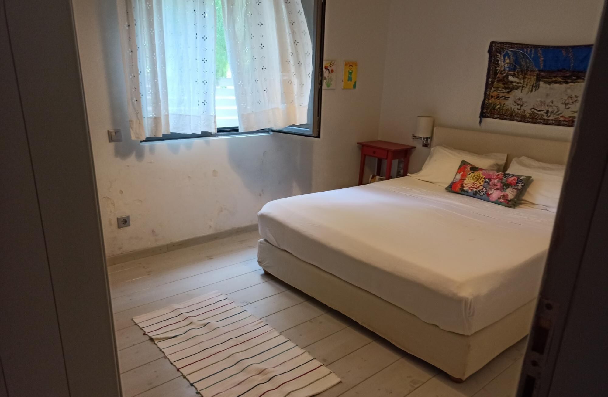 A bedroom with a bed, a window with curtains, a bedside table, and a wall-mounted lamp.
