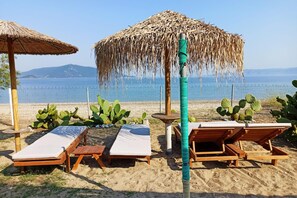 Zona de relax a la platja amb gandules de fusta i paraigües de palla, una planta de cactus i una vista al mar i a les muntanyes de fons.