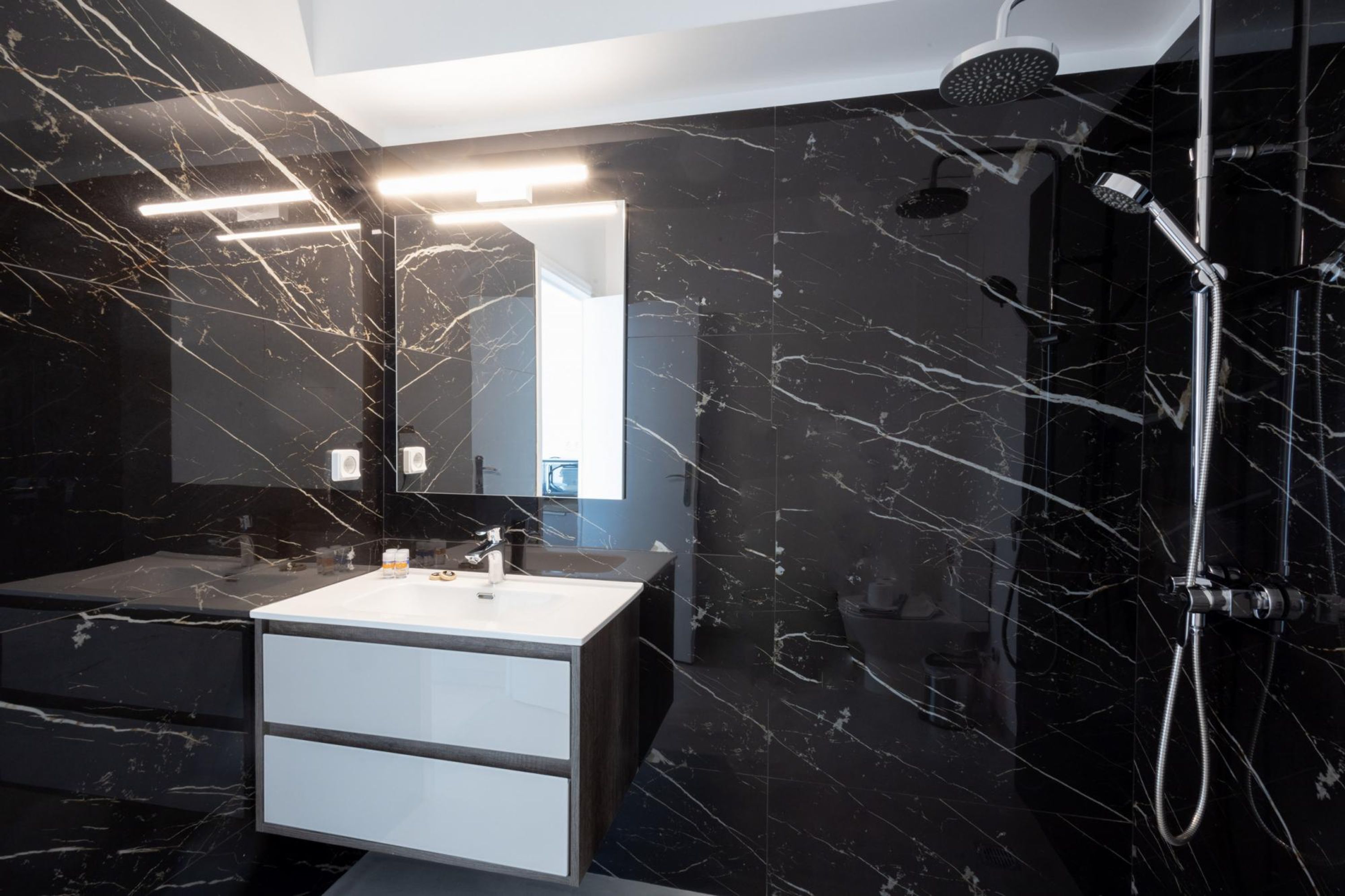 Un baño moderno con piso de mármol, lavamanos blanco, un espejo grande y una ducha con múltiples cabezales.