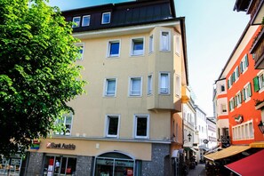 En byggnad med flera våningar, med en skylt som visar texten ”Bank Austria”, ett träd framför och en smal gata med butiker.