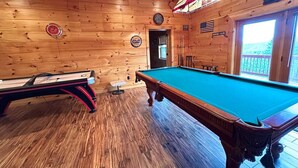 Une table de billard avec une queue de billard et une autre table de billard dans une pièce en bois, où l’on aperçoit une horloge et un panneau accroché au mur.