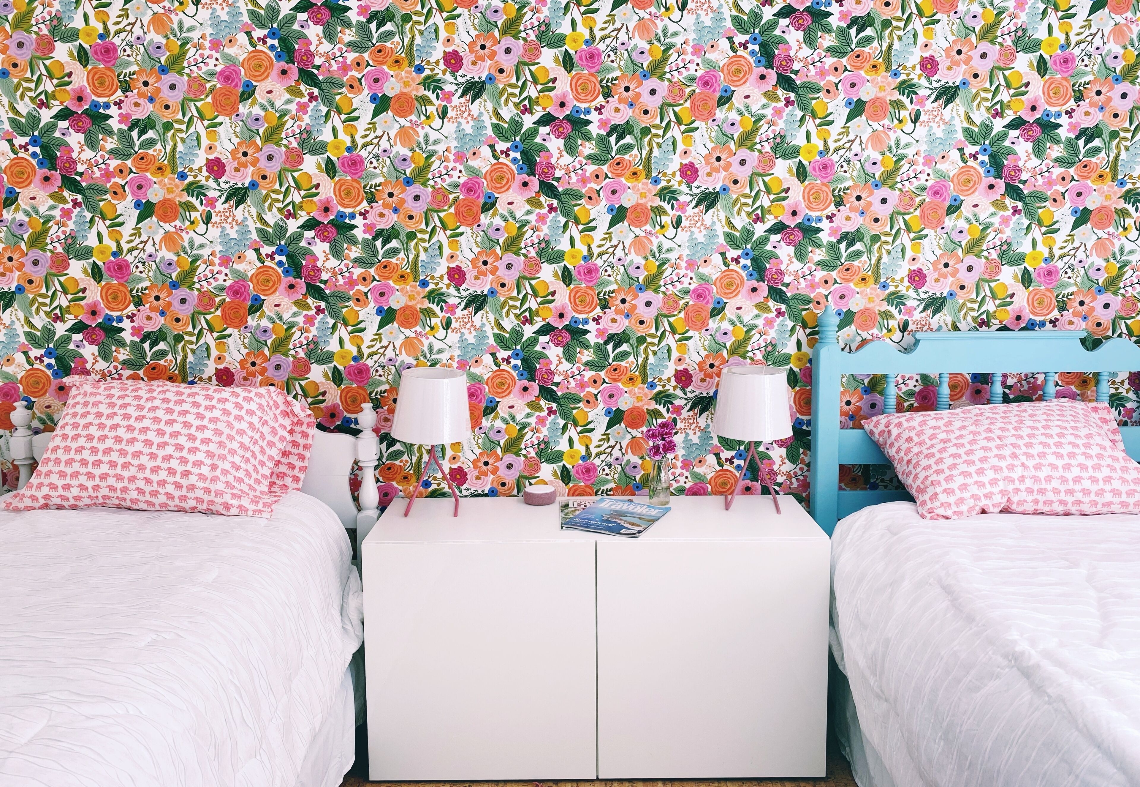 A bedroom with two beds, a white bedside table with lamps, and a floral wallpaper.