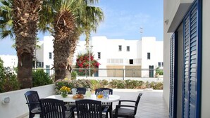 A terrace with a table and chairs, palm trees, and a white building in the background. - Villa Julie in Protaras (Paralimni)