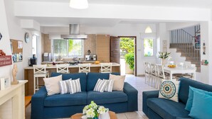 A spacious living area with a blue sofa set, a coffee table with flowers, and a kitchen in the background. - Protaras Villa Athamas Blue (Paralimni)