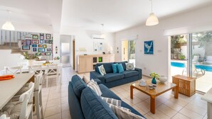 A spacious living area with a blue sofa, a wooden coffee table, and a dining table with chairs. There is a kitchen area with a white countertop and a dining table. A large window provides a view of a swimming pool. - Protaras Villa Athamas Blue (Paralimni)