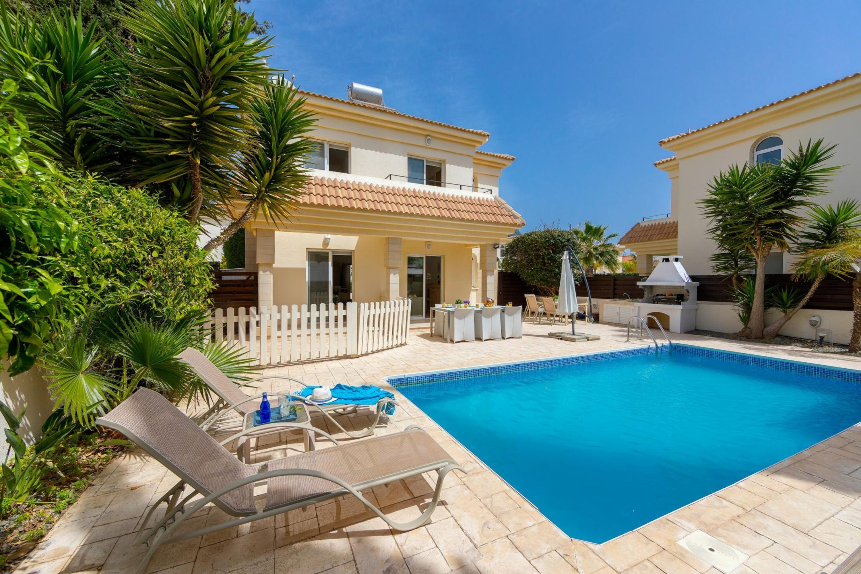 A swimming pool with lounge chairs and a patio area, surrounded by a white picket fence and palm trees.