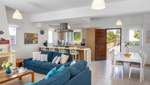 A spacious living area with a blue sofa, a dining table with chairs, a kitchen area, and a view of the outdoors. - Protaras Villa Athamas Blue (Paralimni)