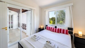 A bedroom with a bed, a nightstand, a window with curtains, and a sliding door leading to a balcony. - Cape Greco Palm Trees Villa (Ayia Napa)