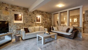 A living room with stone walls, a white sofa, a coffee table, and a TV. - Palmrise Luxury Villas - Anemos Villa (Kassandra)