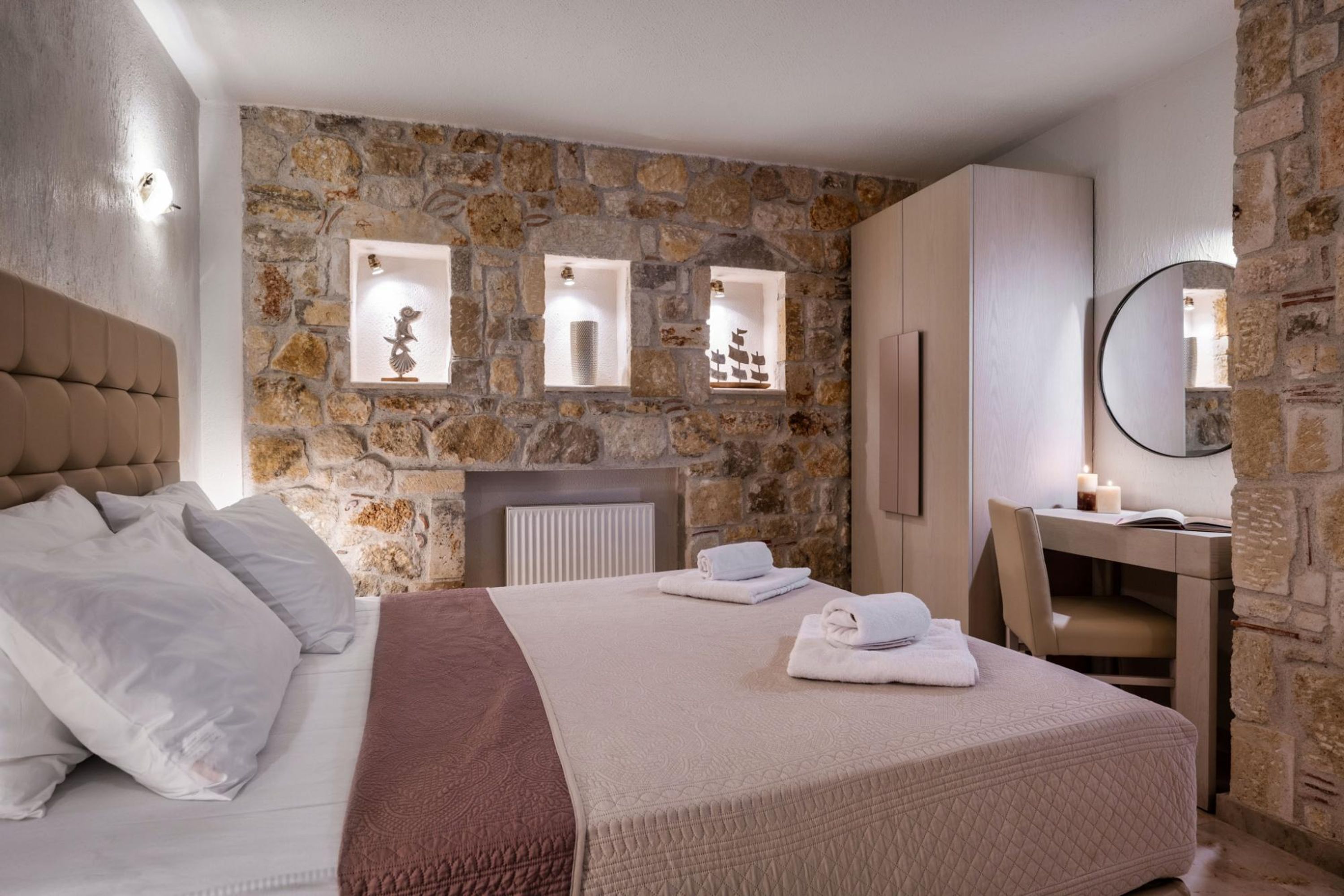 Un dormitorio con pared de piedra, cama con ropa blanca, escritorio con silla y espejo.