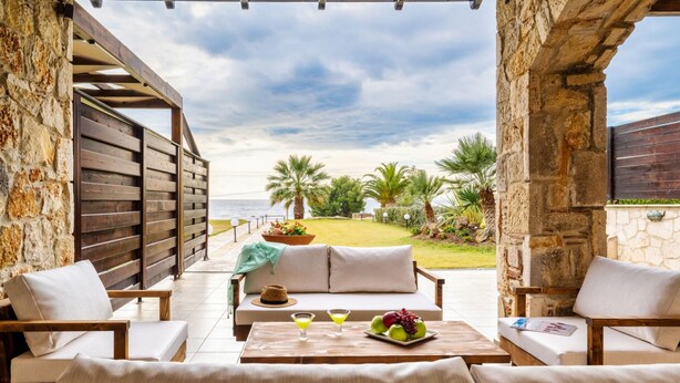 A patio with a wooden table, two sofas, and a stone wall. - Palmrise Luxury Villas - Anemos Villa (Kassandra)