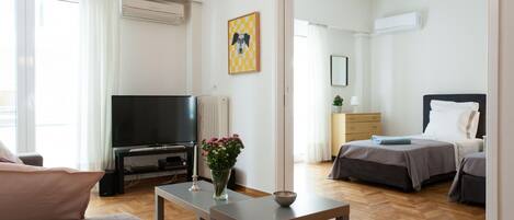 A modern hotel room with a TV, a bedside table with a vase of flowers, a bed with a headboard, and a wooden floor.