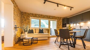 A modern living room with a sofa, a dining table with chairs, a coffee table, and a kitchen area with cabinets and a window. - Tevini Alpine Apartments - Schmittenblick (Zell am See)