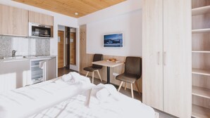A modern hotel room with a wooden ceiling, a kitchenette, a bed, a small table with chairs, and a wardrobe. - Appartements Sulzer - TOP 23 (Zell am See)