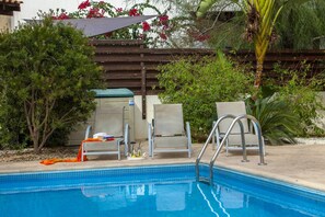 Une piscine avec deux chaises longues et une petite table, entourée de verdure et d’une clôture en bois.