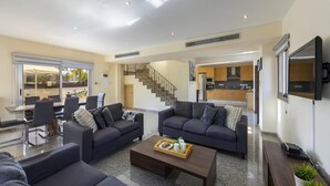 A modern living room with dark sofas, a coffee table, and a dining area with chairs and a table. A kitchen is visible in the background with wooden cabinets and a TV mounted on the wall. - Theodora 1 in Protaras (Paralimni)