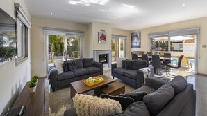 A spacious living room with two sofas, a coffee table, and a fireplace. There is a dining area with a table and chairs, and a pool visible through the glass doors. - Theodora 1 in Protaras (Paralimni)