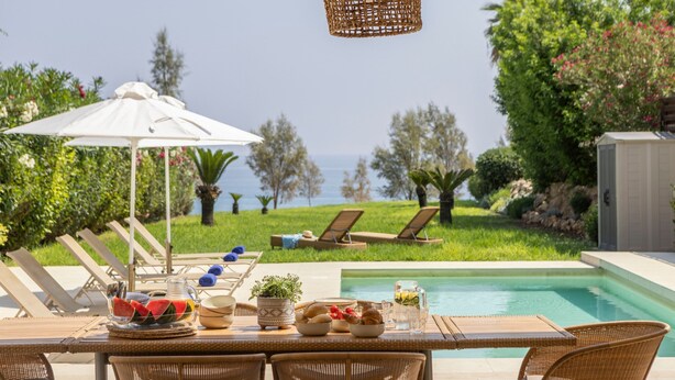 A poolside dining area with wicker furniture, a table set with plates, bowls, and a pitcher, and a view of the sea. - Protaras Beachfront Villa Pietra (Paralimni)