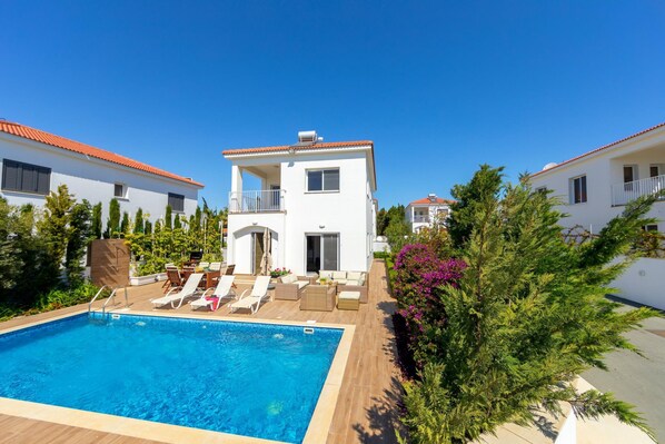 A poolside area with a rectangular pool, lounge chairs, and a white building with a balcony. - Napa Bay View Villa 2 (Ayia Napa)