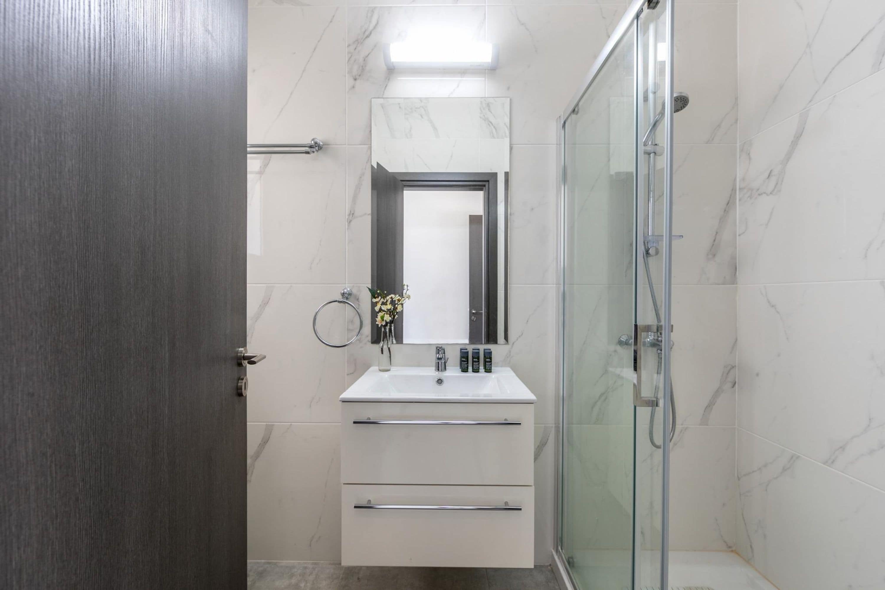 Un baño moderno con cabina de ducha de vidrio, un lavamanos blanco con encimera de mármol y una puerta de madera.