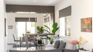 Een moderne woonkamer met een grijze bank, een eethoek met een tafel en stoelen, en een groot raam met uitzicht op bomen.
