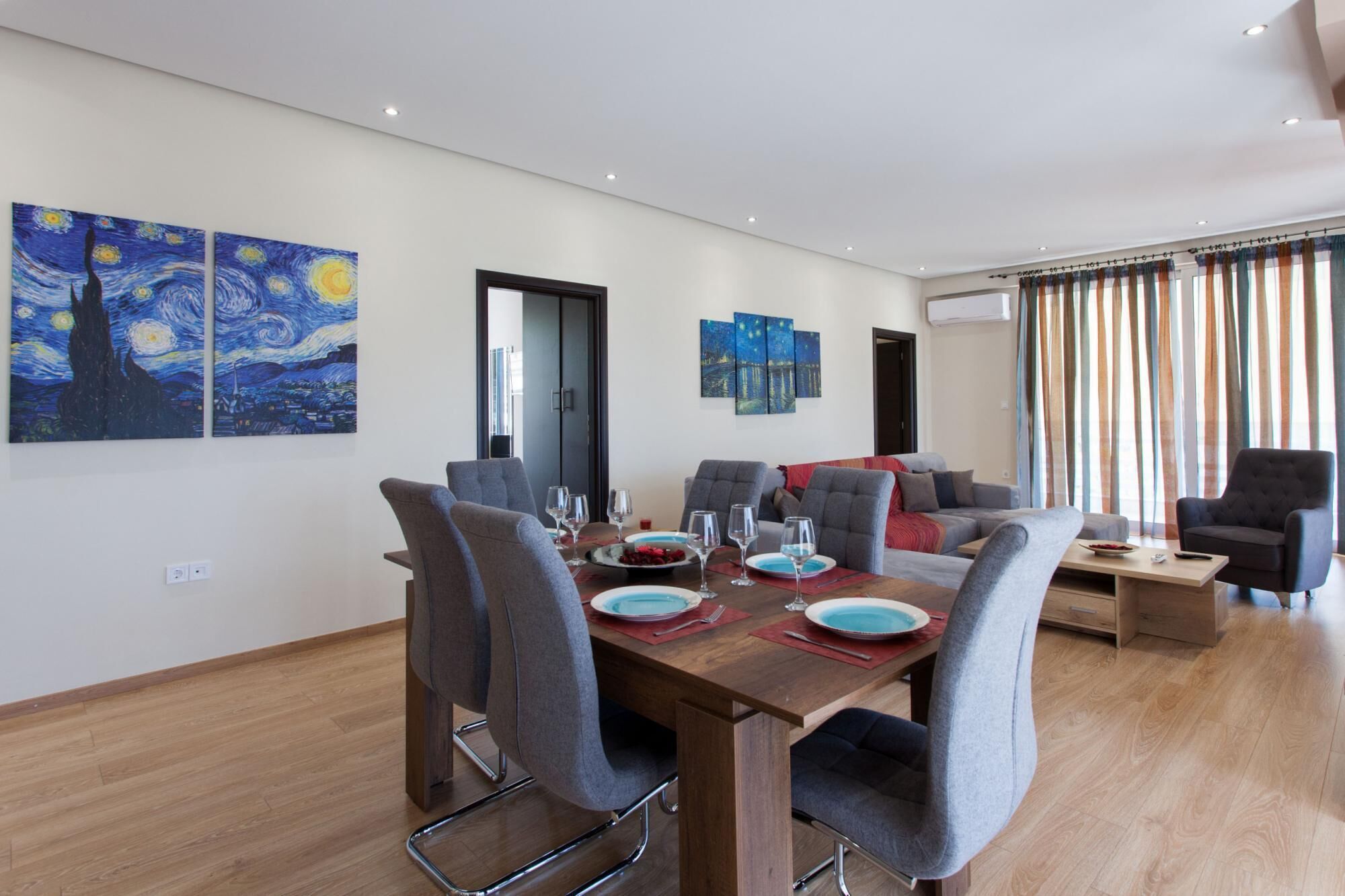 Een moderne eetruimte met een houten tafel voor vier personen, omringd door grijze bekleden stoelen. Er hangen drie schilderijen aan de muur, en een bank met kussens is op de achtergrond te zien.