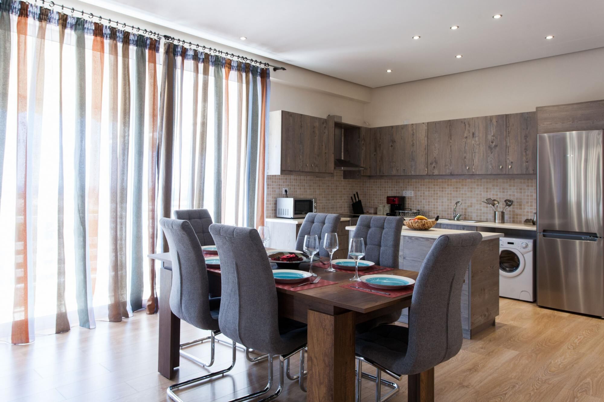A modern kitchen with wooden cabinets, a stainless steel refrigerator, and a dining area with a wooden table and grey chairs.