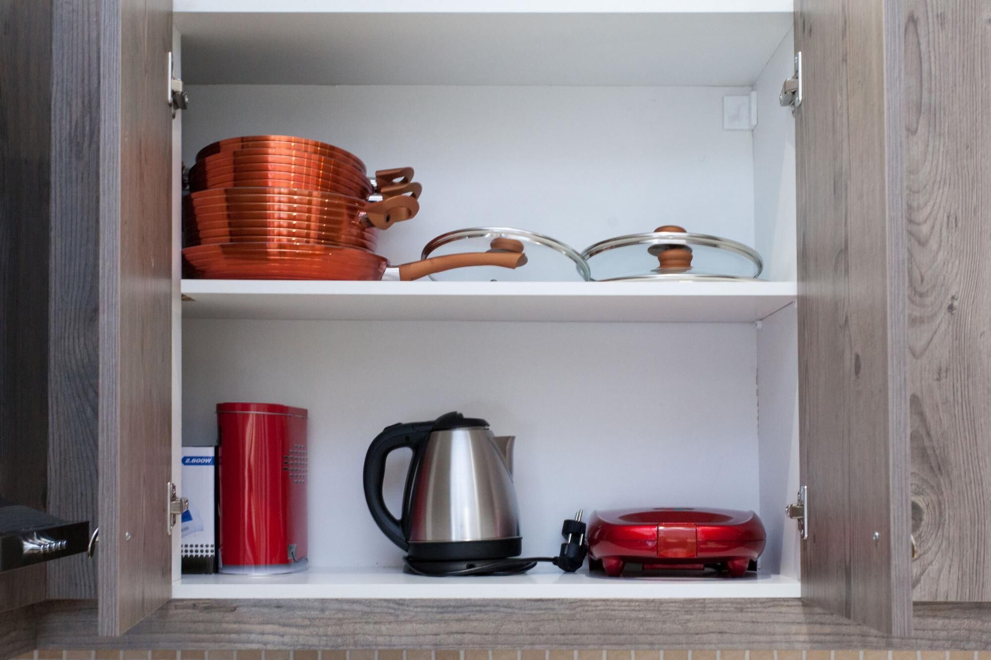 A kitchen cabinet with a microwave, kettle, and red mug on the lower shelf, and copper cookware on the upper shelf.