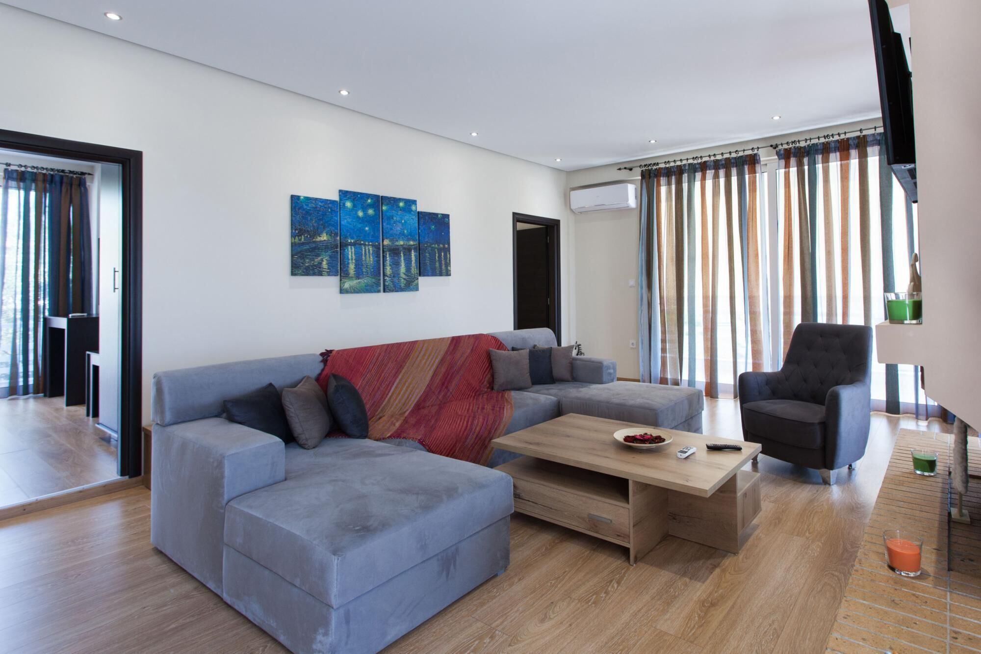 Una sala de estar moderna con un sofá seccional gris, una mesa de centro de madera y un sillón negro. Hay un cuadro en la pared y un equipo de aire acondicionado montado en la pared.