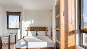 A modern bedroom with a wooden headboard, a bed with white linens, a desk with a chair, and a view of buildings through a window. - Explore Nightlife Near an Apartment With an Acropolis View (Athens)