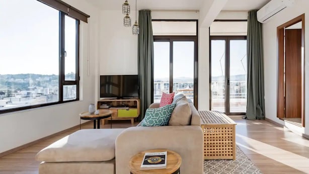A modern living room with a large window, a sofa, a TV, and a small table. - Explore Nightlife Near an Apartment With an Acropolis View (Athens)