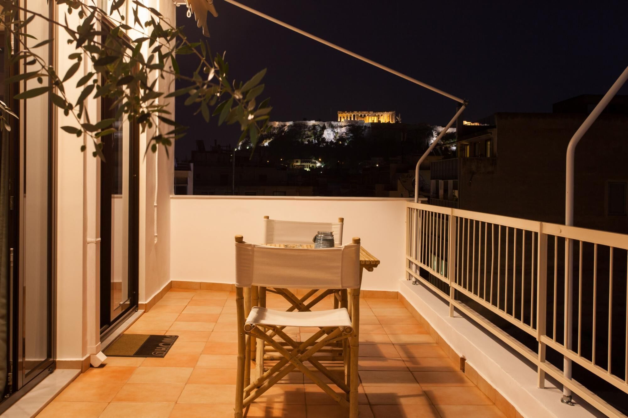 A balcony with a chair and a table, overlooking a cityscape at night with a building illuminated in the background.