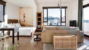 A modern hotel room with a bed, a sofa, a small table, a TV, and a large window offering a city view. - Explore Nightlife Near an Apartment With an Acropolis View (Athens)