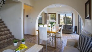 A spacious living area with a dining table, chairs, and a sofa. There is a staircase on the left and a large window providing natural light. - Villa Buena Vista in Protaras (Paralimni)