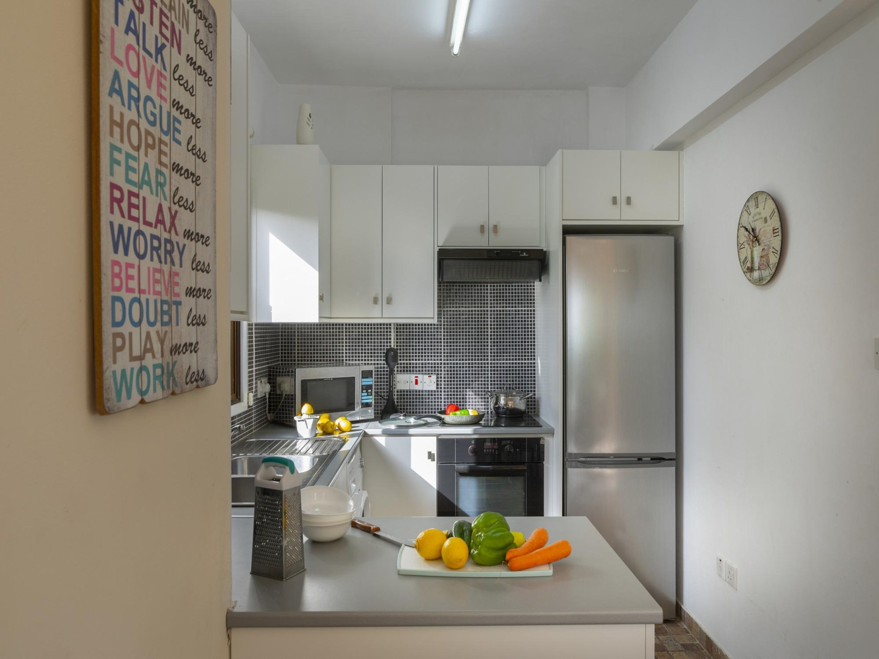 A modern kitchen with white cabinets, a black tiled backsplash, a stainless steel refrigerator, and a word cloud on the wall.