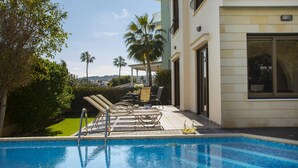 A swimming pool with lounge chairs and a table, surrounded by greenery and a building with a balcony. - Villa Buena Vista in Protaras (Paralimni)