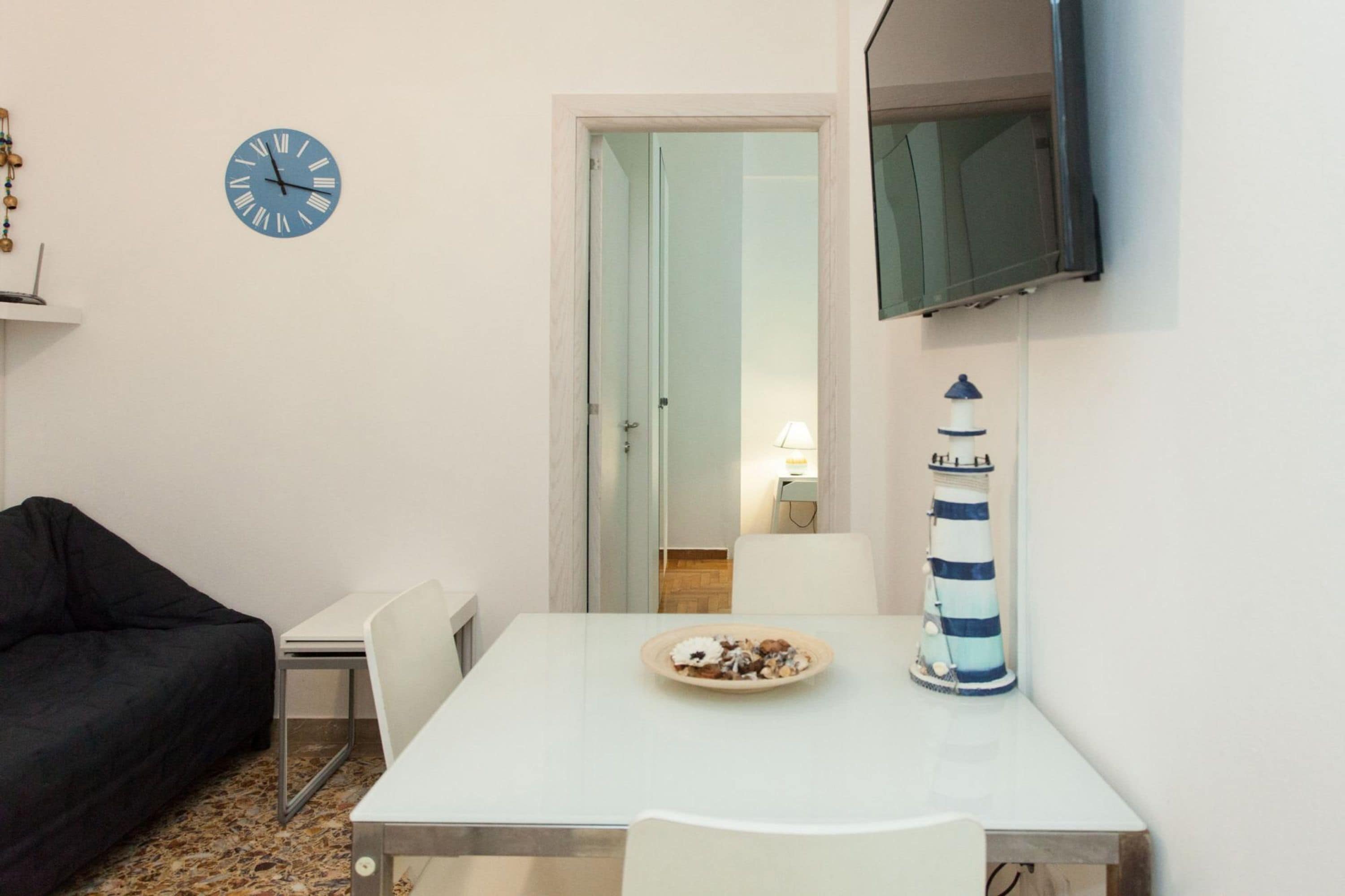 A room with a white table, a black couch, a clock, a television, and a decorative lighthouse.