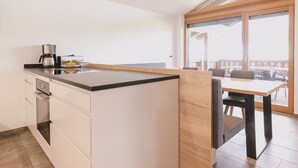A modern kitchen with a central island, a dining area with a table and chairs, and a large sliding glass door leading to an outdoor patio. - First IN Mountain Chalet B (Kaprun)